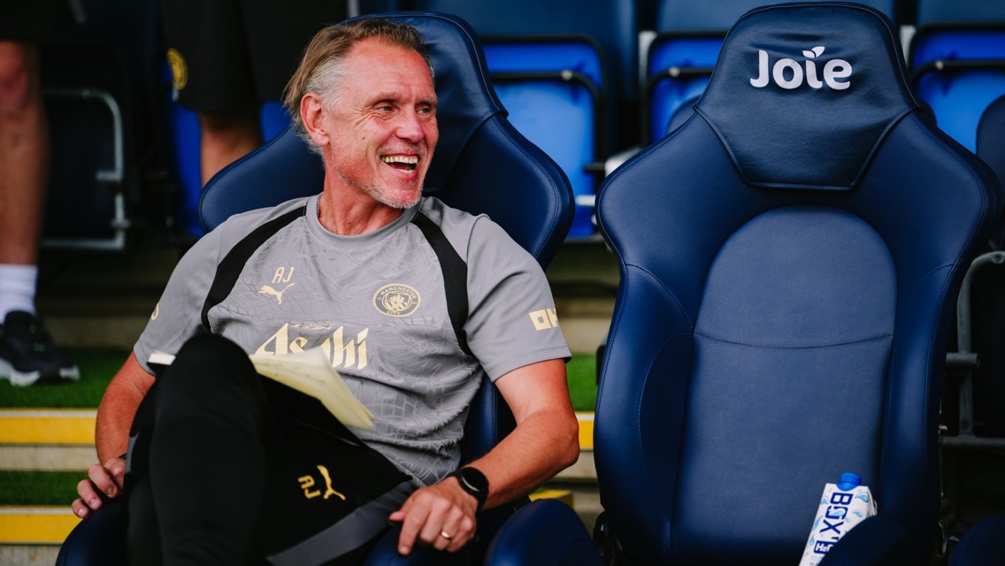A man wearing Manchester City sports gear is sitting on a chair at a stadium, smiling and holding a notebook. The chair next to him has the word 'Joie' on it.